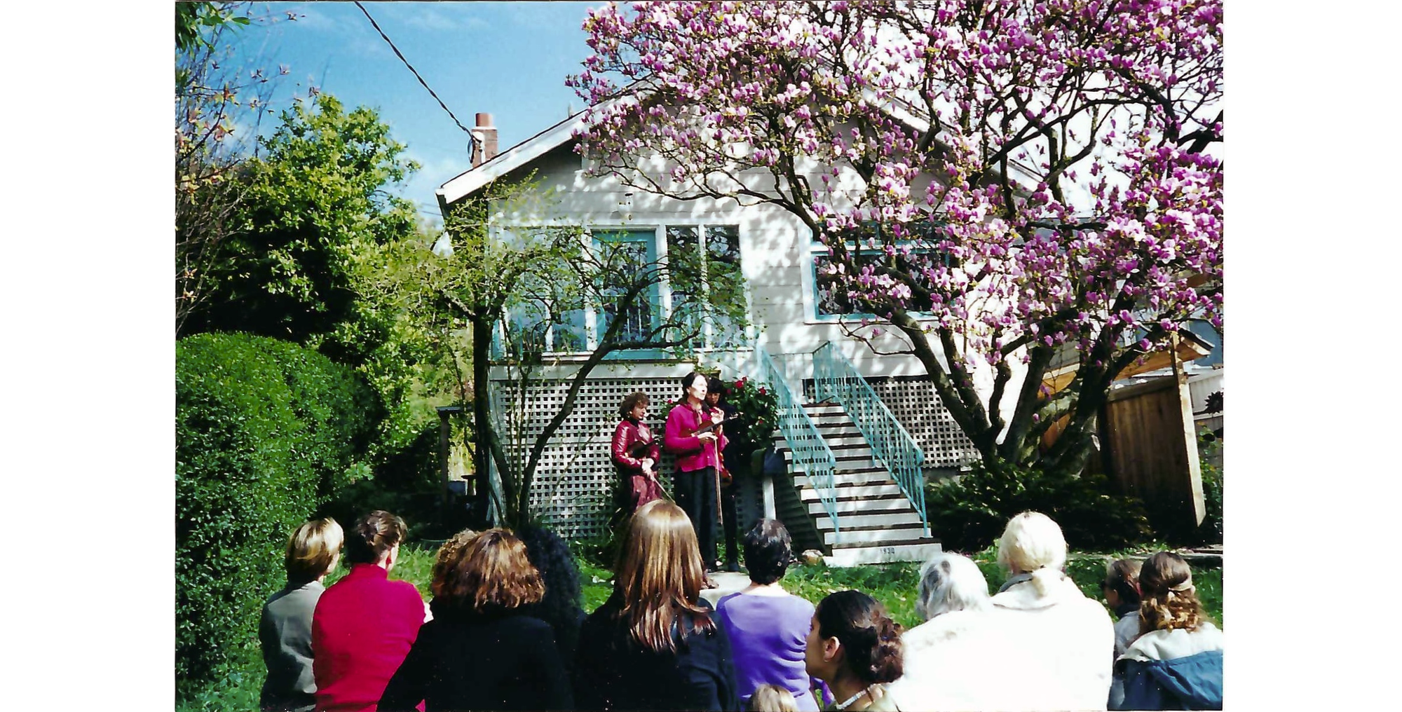 Photo of two women wit violins stand in front of house with magnolia in bloom, with group of women in the foreground