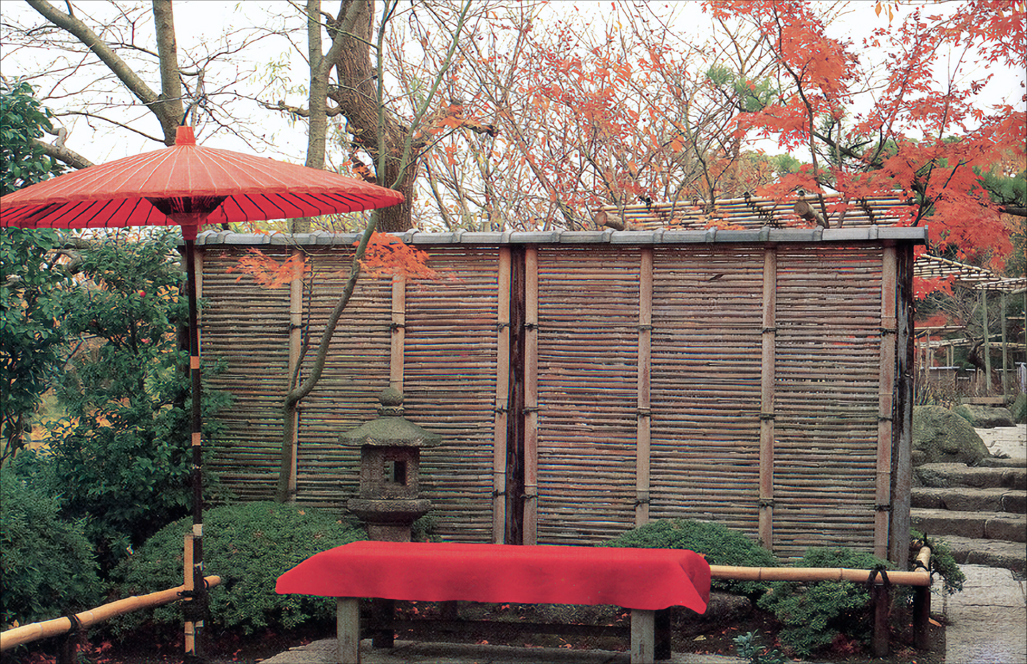 Photo of a bamboo screen with a a red Chinese umbrella atop a pole on the left, and a bench with a red cover in front