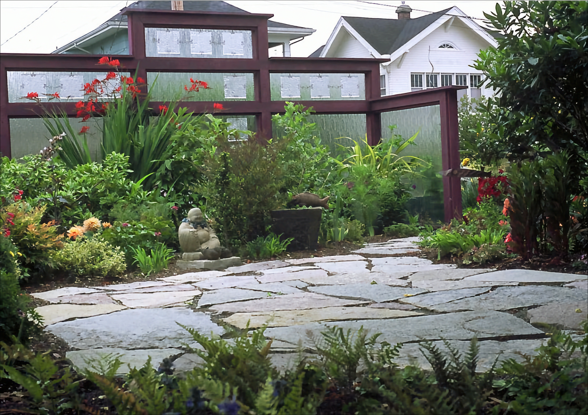 Photo of several panels of tempered glass frame by dark wook, behind a small gardem with red crocosmia and green plantings behind as stone Buddha statue, all facing a paved stone patio