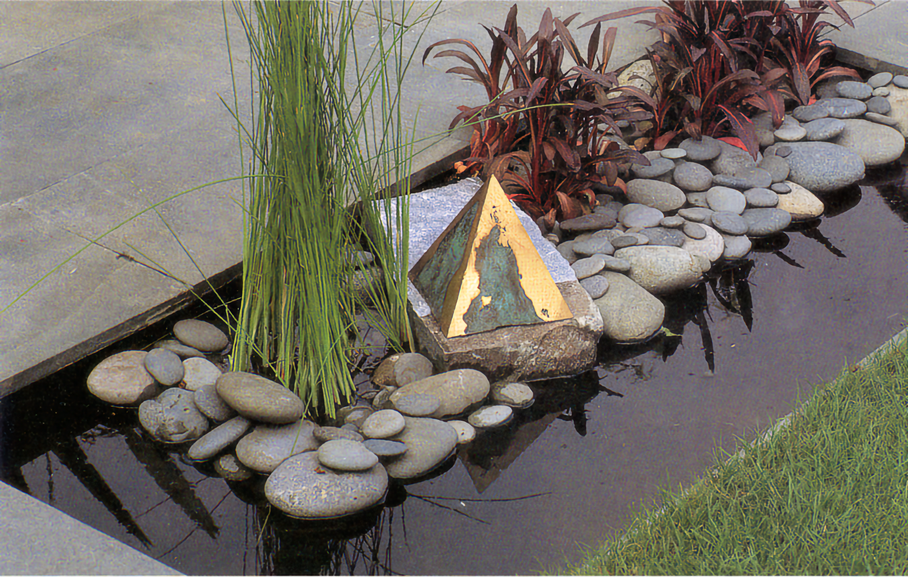 Photo of small rectangular pool with round stones underneath green and red plants and a small metal pyramid