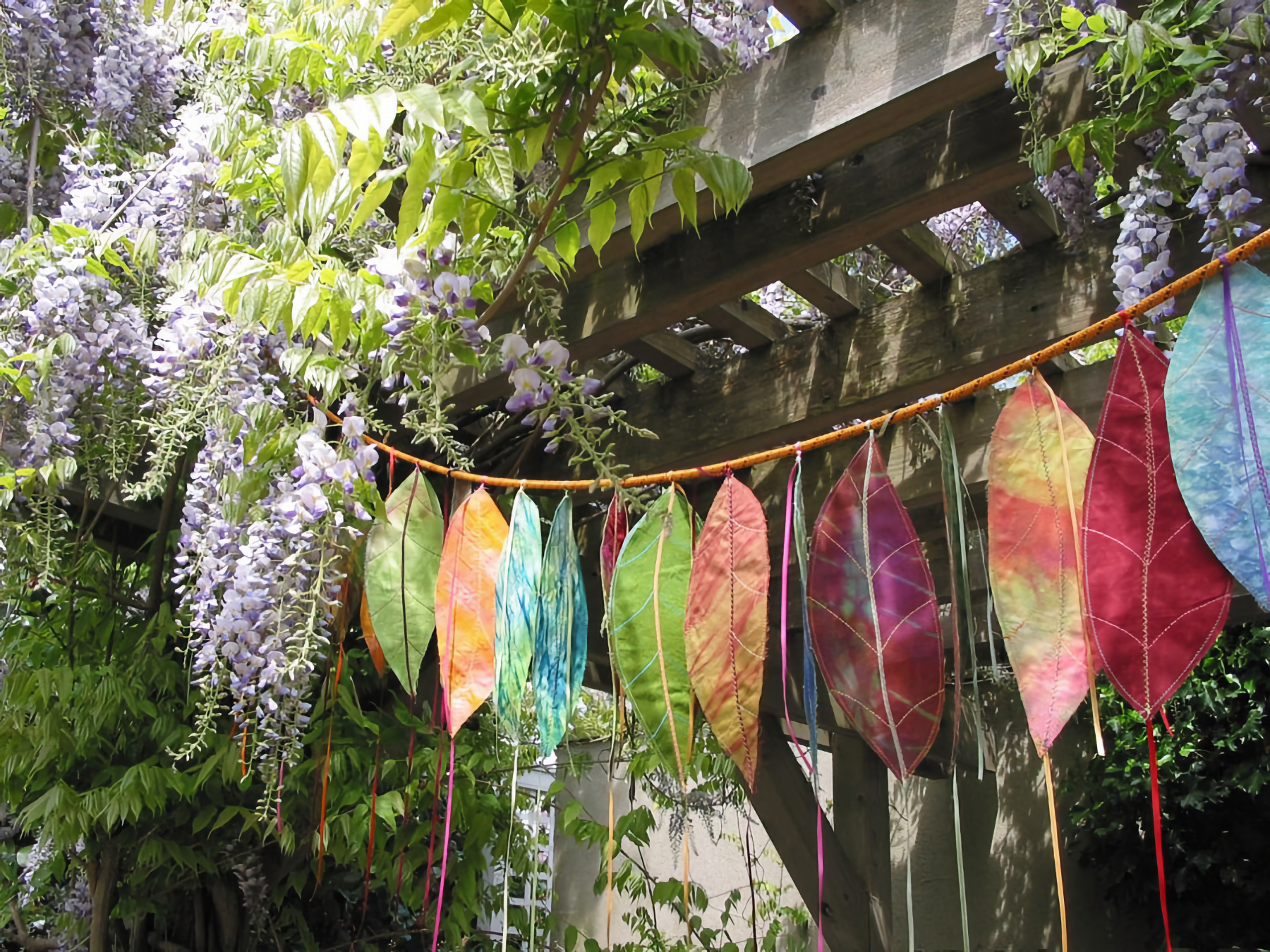 Photo of purple wisteria on arbor in back, with multi-colored cloth leaves on cable in front