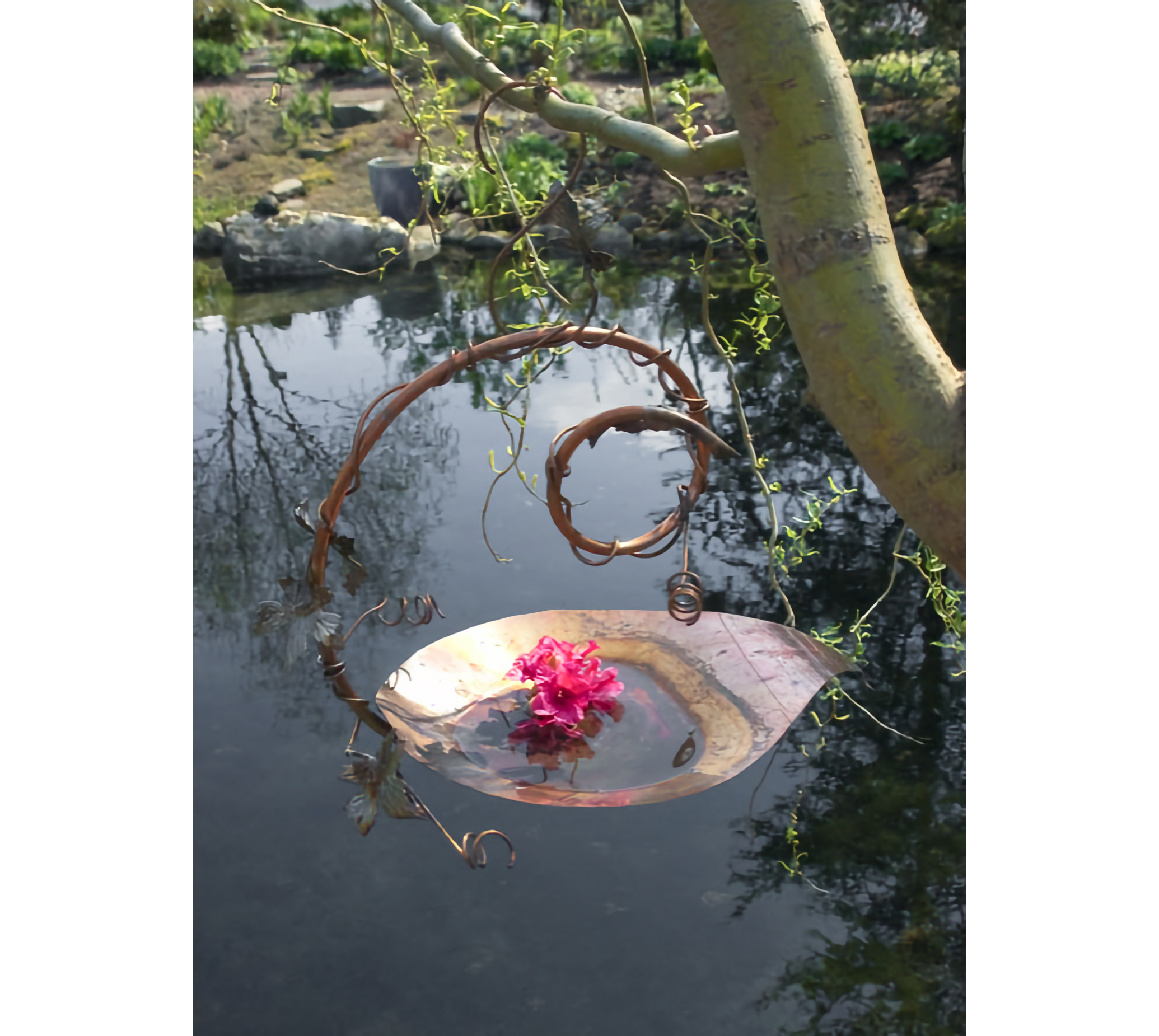 Copper birdbath with spiral tom and red flower in basin, hanging on tree limb above water