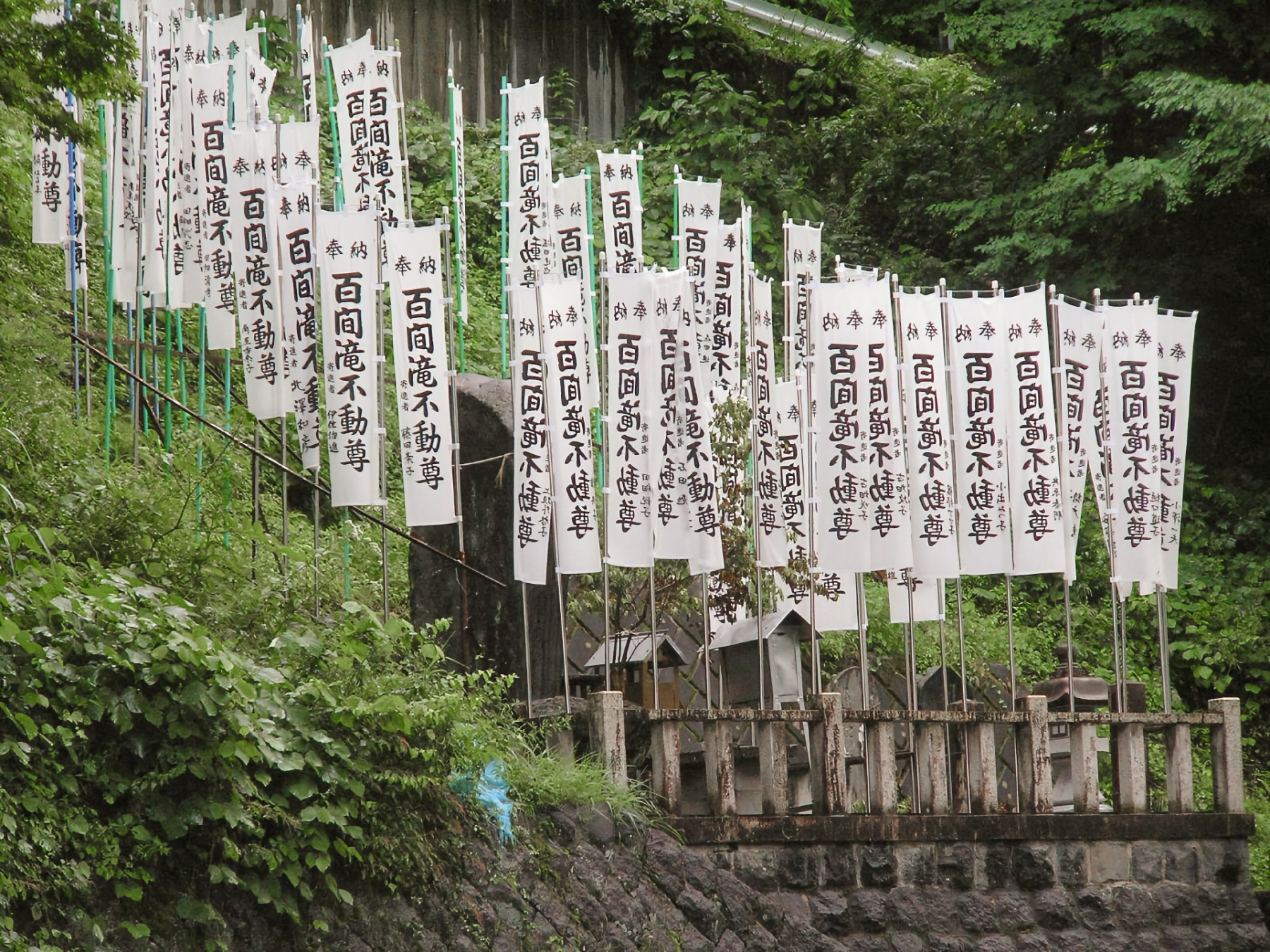 Multiple tall vertical white banners with Japanese characters, on side of a hill in front of entrance to a shrine