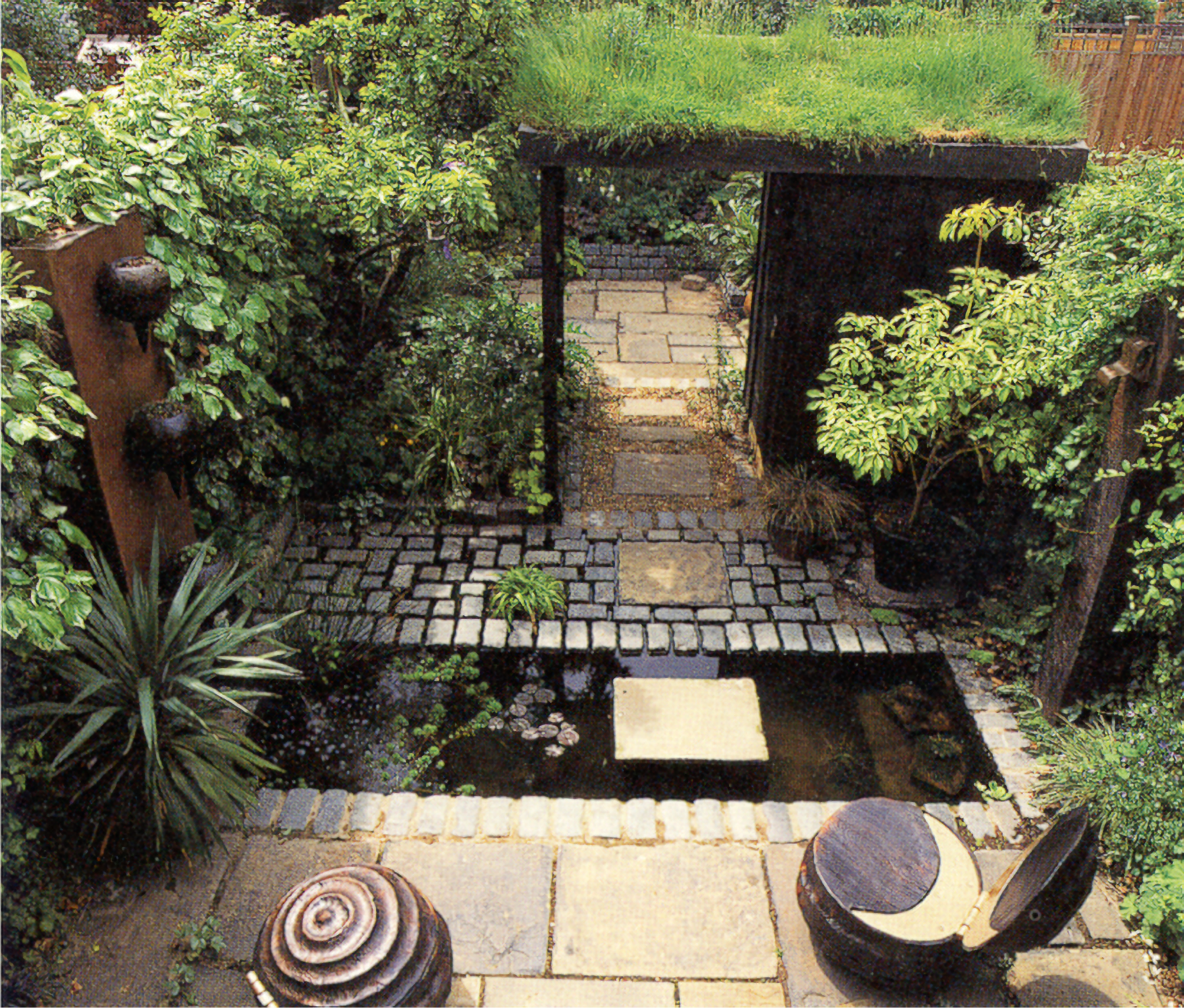 Photo of a grass-thatched garden entry past a reflecting pool with paving stone bridge and shrubs on the sides