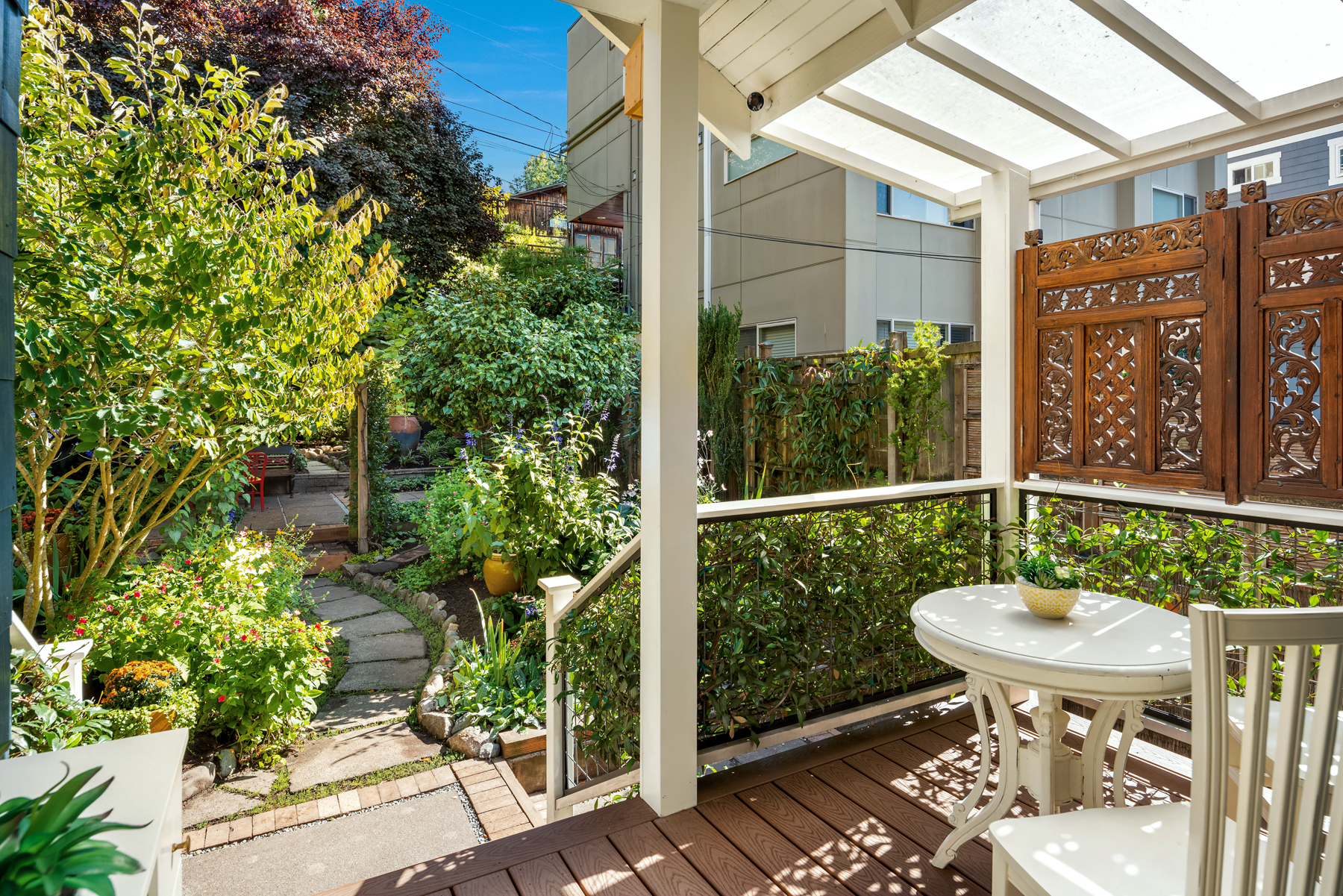 Photo of small coverd back deck with white table, Indonesian wooden screen on right deck railing, jasmine on the bottom right and front railing, looking out over a garden