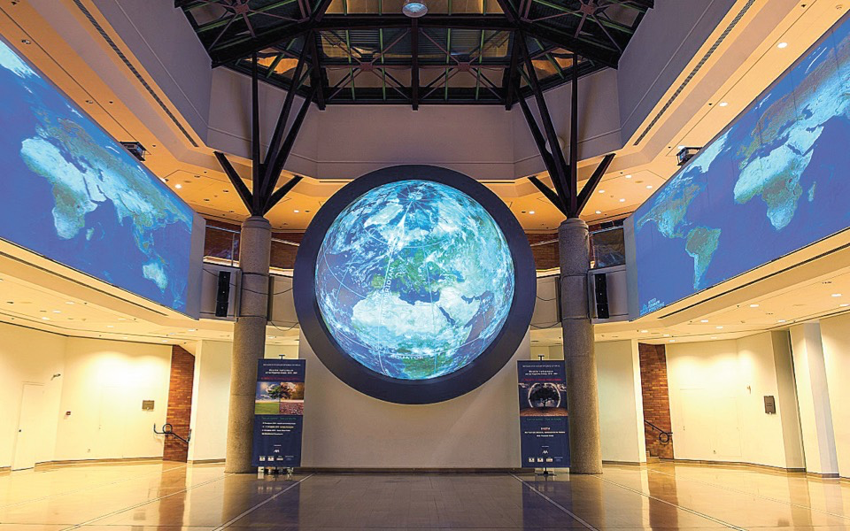 Photo of large room with maps of earth on side walls, and dome of earth in center, between two columns