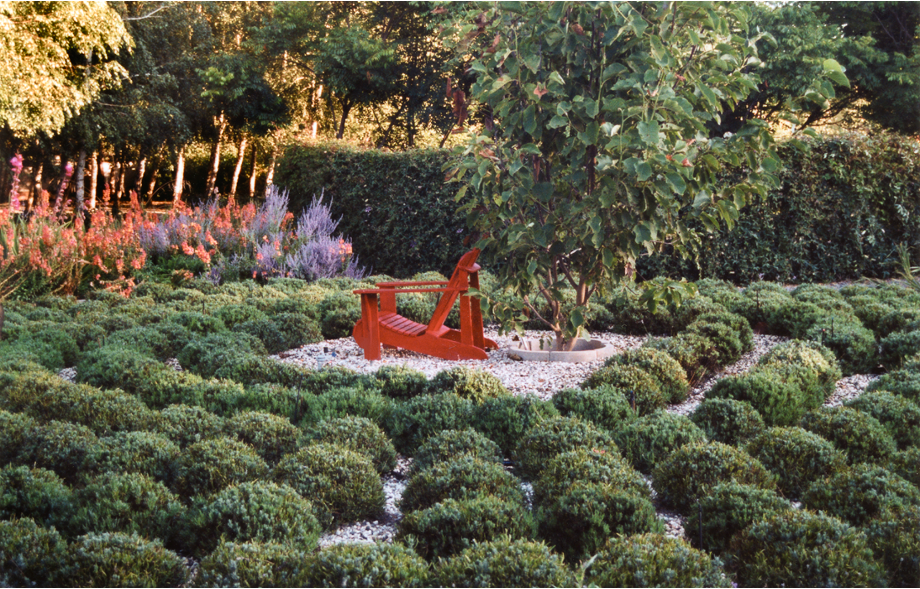 A wooden chair in the center next to a small tree is surrounded by low lavender plants, with lavender flowers and salmon blossoms to the left, all in front of a high shrub border