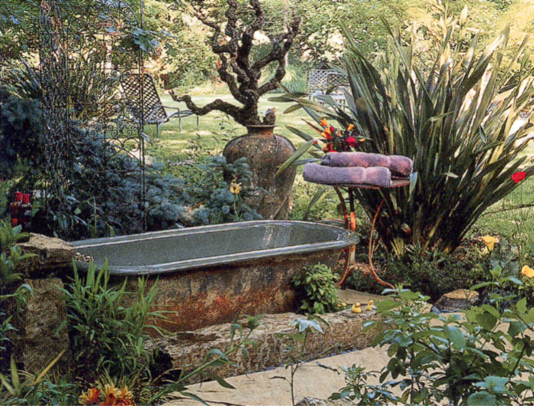 Photo of a blue tub on a stone riser, surrounded by elaborate plantings, with an iron table with purple towels at the head.