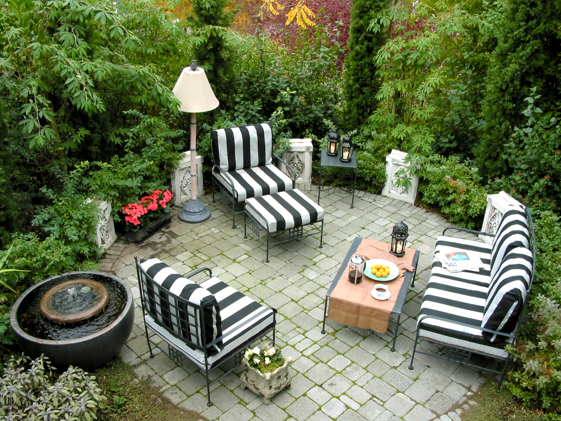 Photo from above looking at three zebra chairs  on a round stone patio, with a low metal table with coffee and fruit in front of one, a fountain to the left, and small red flowers and green plantings around the perimeter