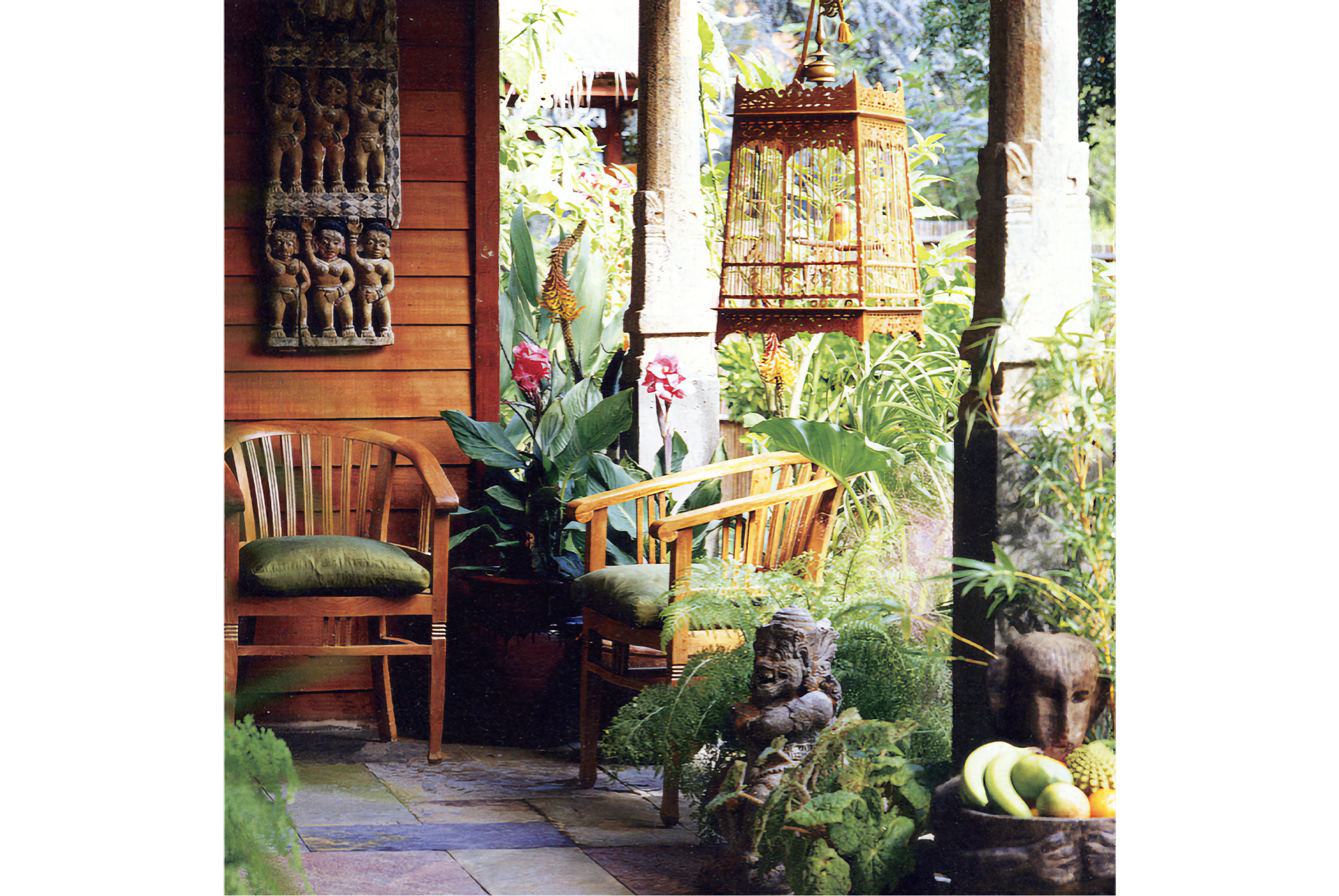 Photo of veranda with two round chairs with green cushions, a wooden bird cage hanging on the right, sculptures, and a lush garden in the background