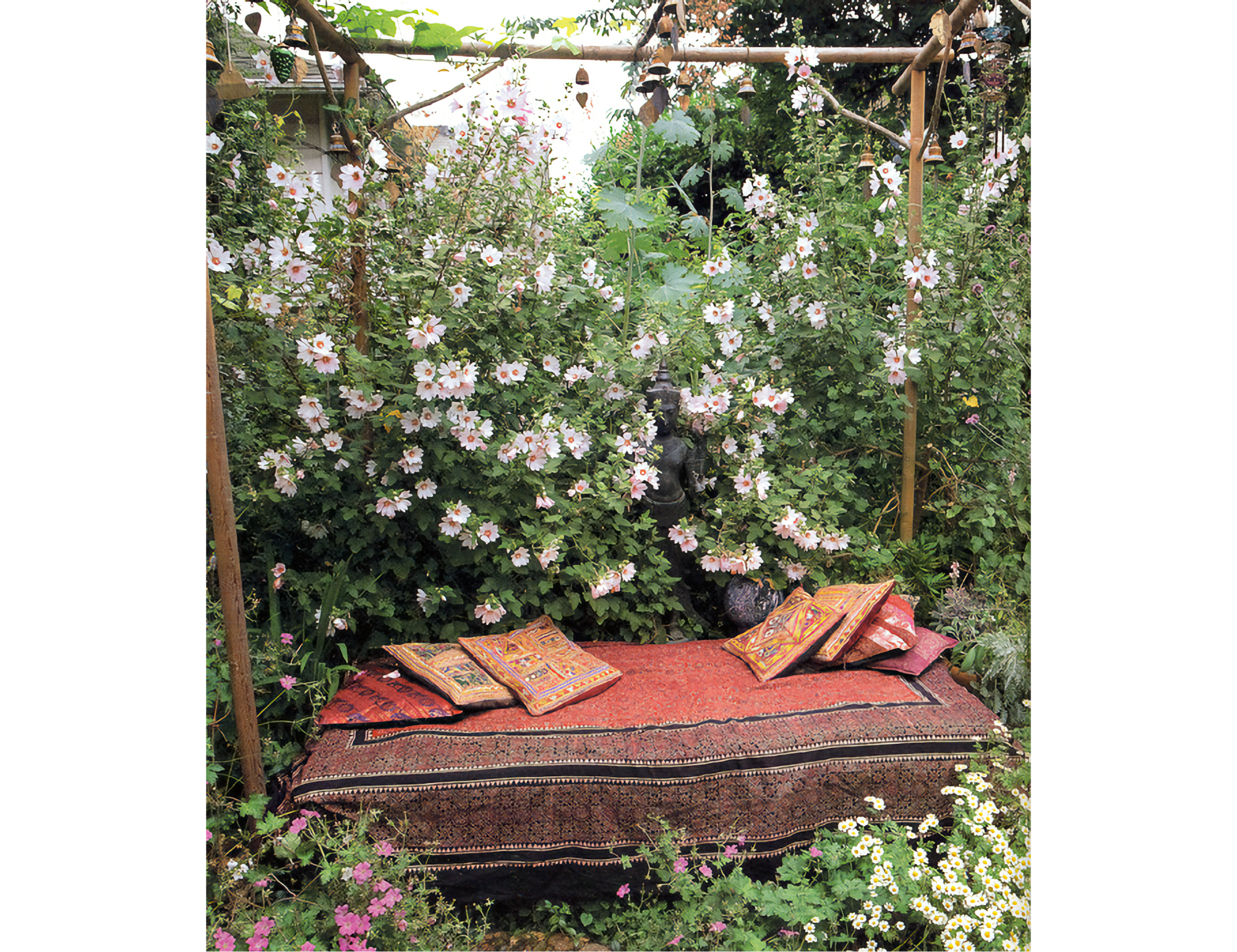 Photo of backless bench covered with red cloth and colorful pillows, underneath a wooden bower with white and red flowers in the back