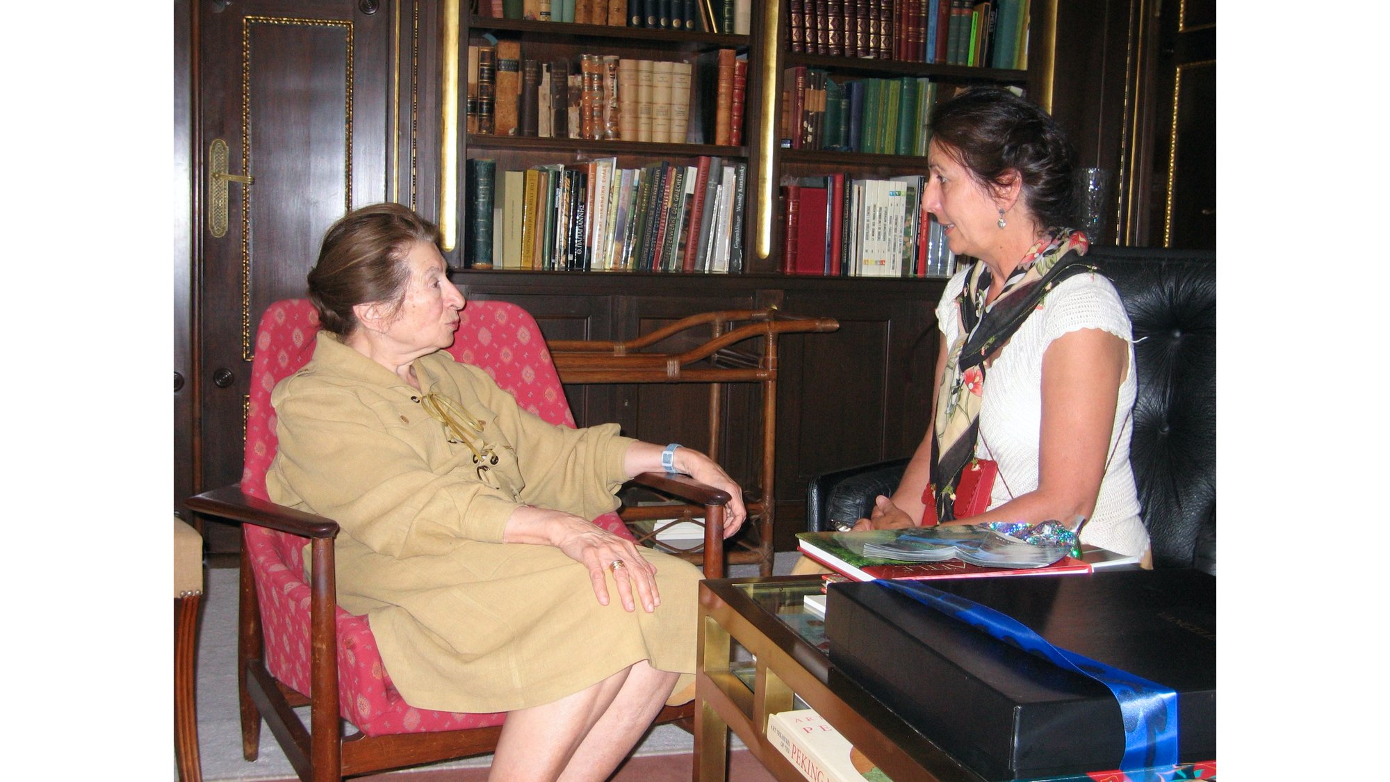 Photo of Niki Goulandris sitting in red chair in brown dress, facing right, looking at Sandra Dean, sitting in black chair wearing white blouse, looking left, with desk in foreground and bookcases in background