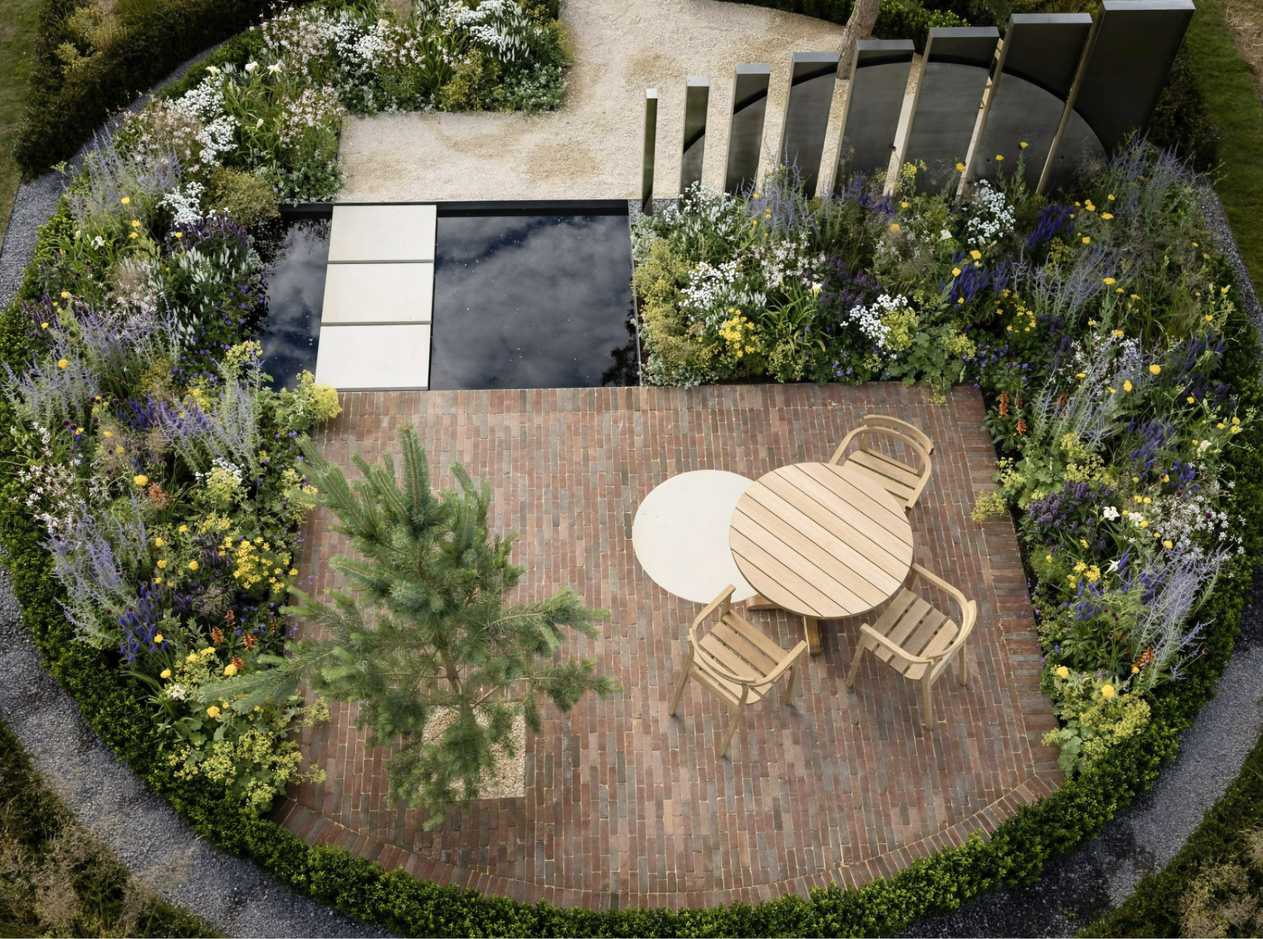 Photo from above of wooden table and four chairs on brick patio with flower plantings spiraling in from left and right, and small square pool in back