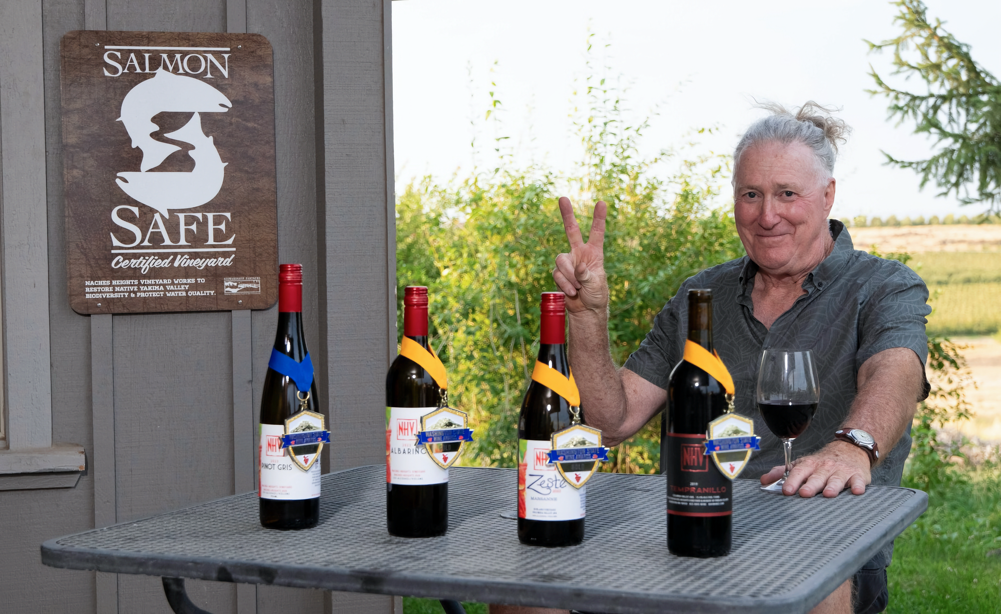 Photo of Phil Cline at outside table with 4 bottles of award-winning wines, smiling and holding up "V" sign with fingers