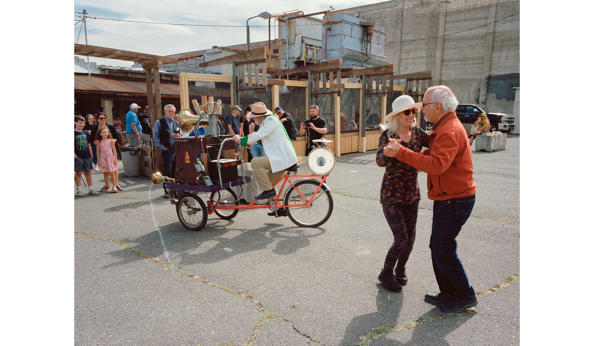 Photo of Trimpin on bicycle playing pump organ, with Sandra and David dancing in front and to right