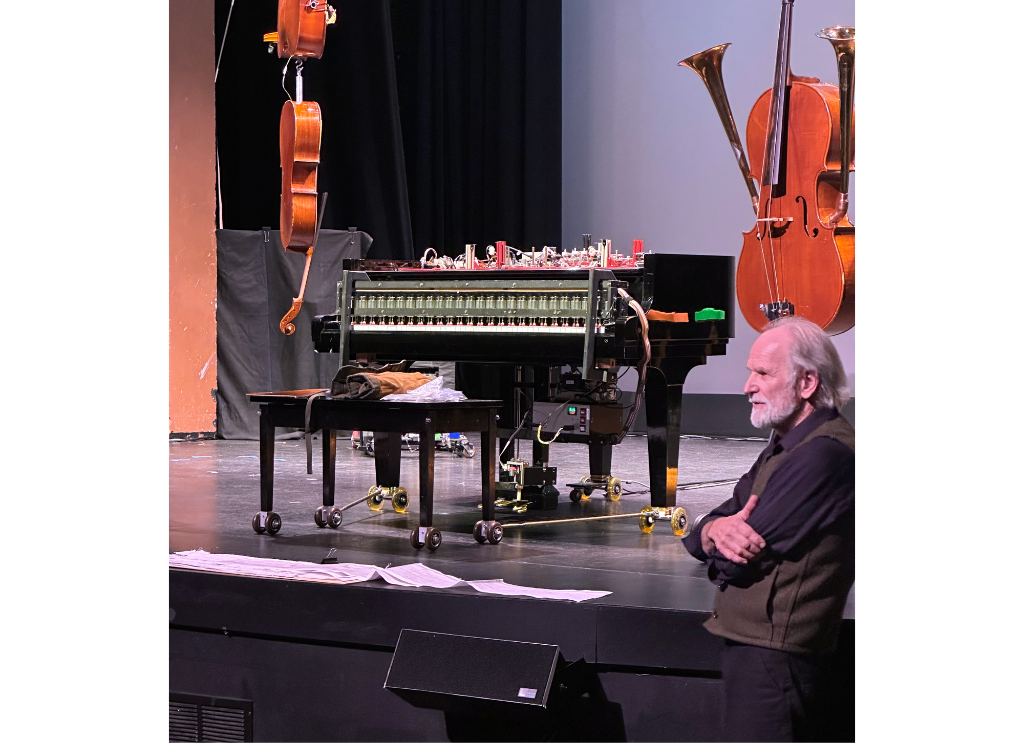 PHoto of Trimpin in front of stage to right, with prepared piano, manuscript, and hanging cellos and horns on stage