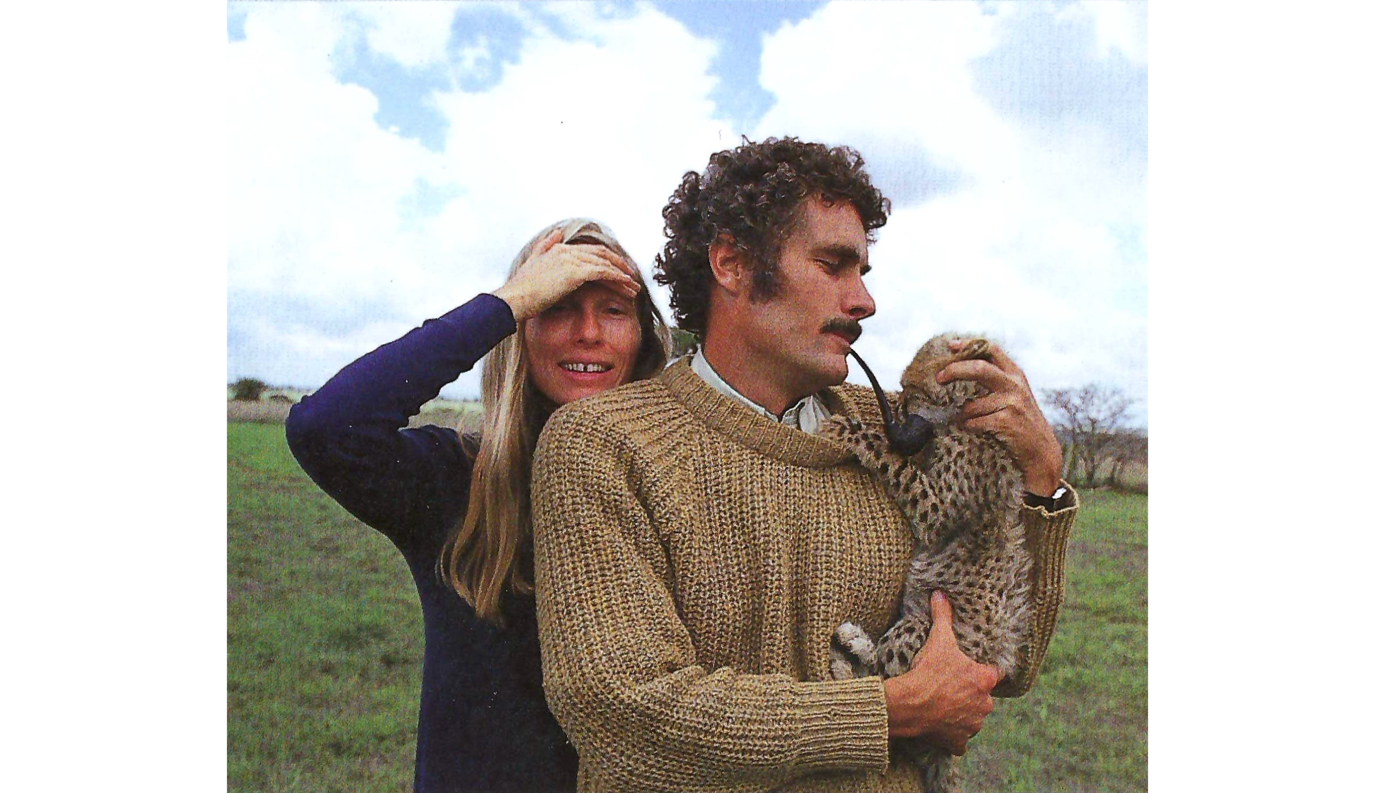 Photo of a man and woman in a field, with the woman in a blue sweater holding her right hand to her forehead as she faces the camera, and the man to her right in a brown sweater and smoking a pipe, looks down to his left at the baby cheetah he is holding in his arms.