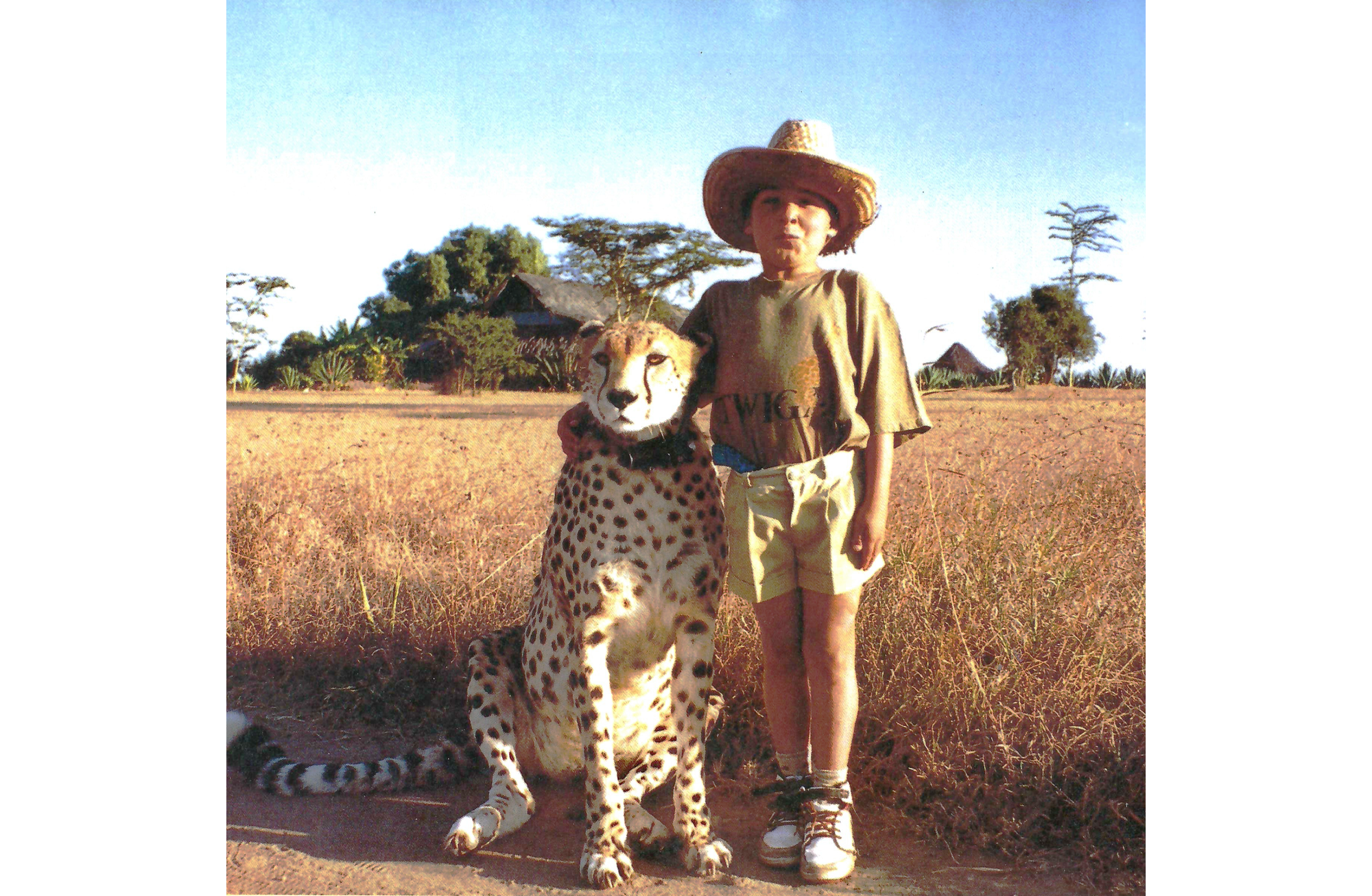 Photo of youn boy standing to right, wearing a straw hat, bwon shirt and shorts, with his right arm around the head of a cheetah seated to his right, both looking at the viewer.