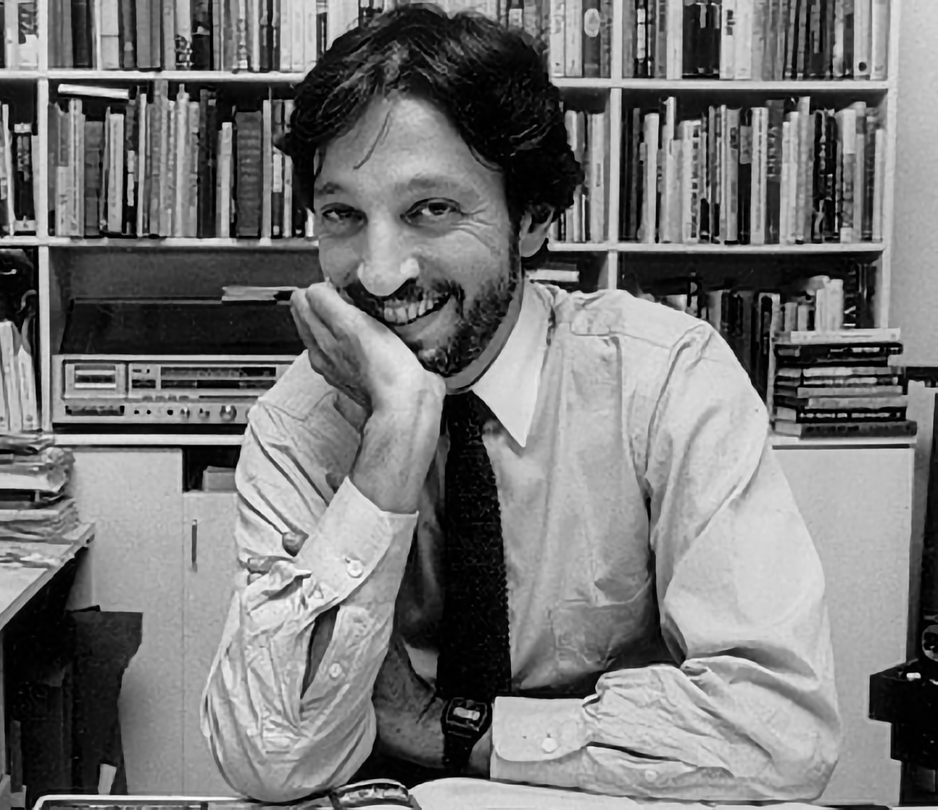 Photo of artist Fred Marcellino in white shirt and necktie, right hand on right cheek, at his desk, smiling and looking directly at camera, with bookshelves in background