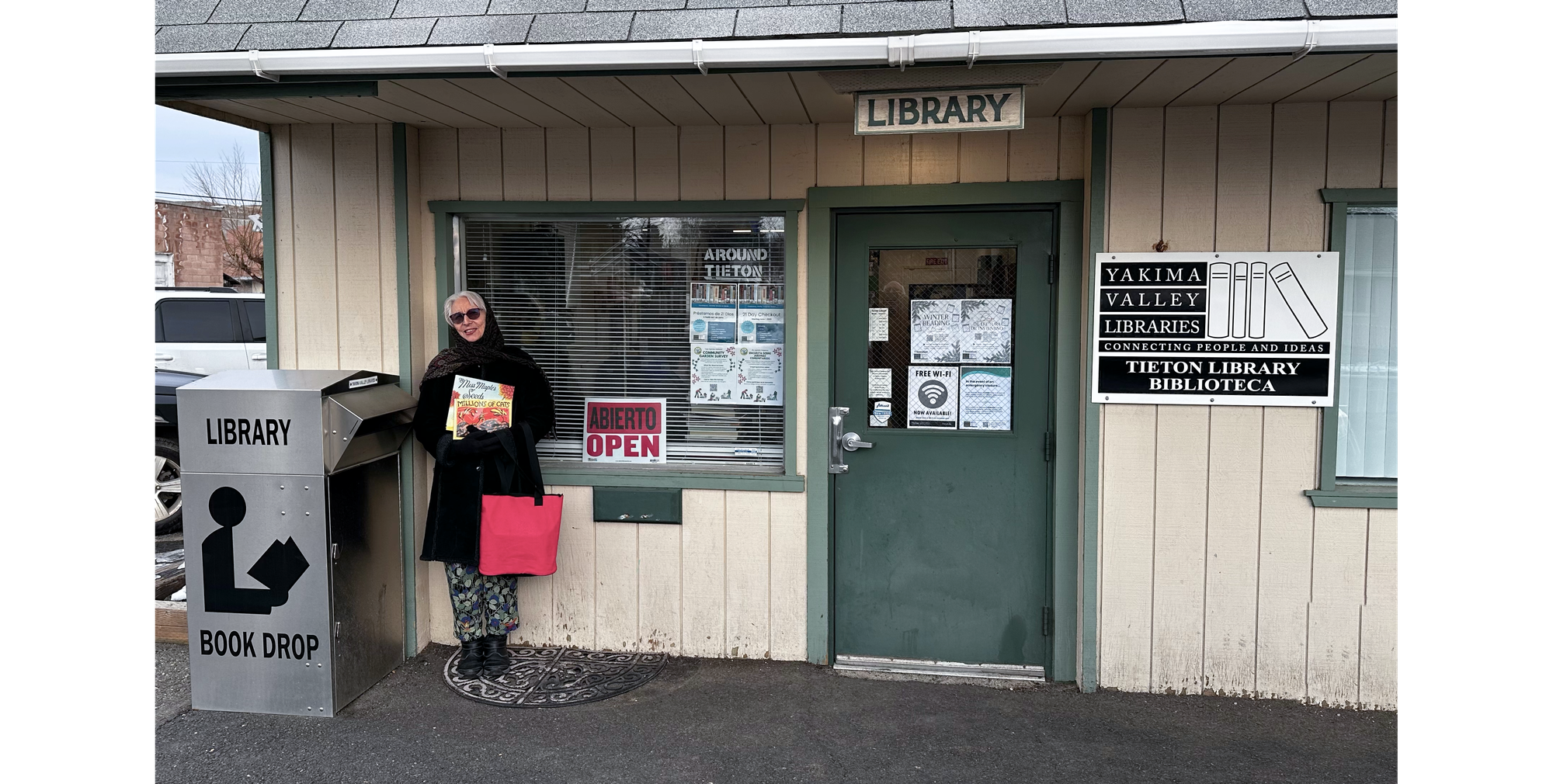 Sandra wearing sunglasses, black coat, and scarf, carrying a red bag and holding two books to her chest while facing the camera, standing outside the Tieton library entrance.