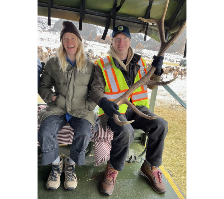 Photo of Anne and Nick Felton in winter clothes sitting on seat in open elk viewing truck, with Nick holding a rack of elk antlers.