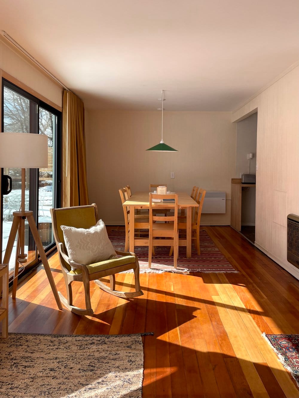 Image of interior of an El Nido cabin, sunlight on wood floor from glass door on left, with chair and pillow, and dining table in background.