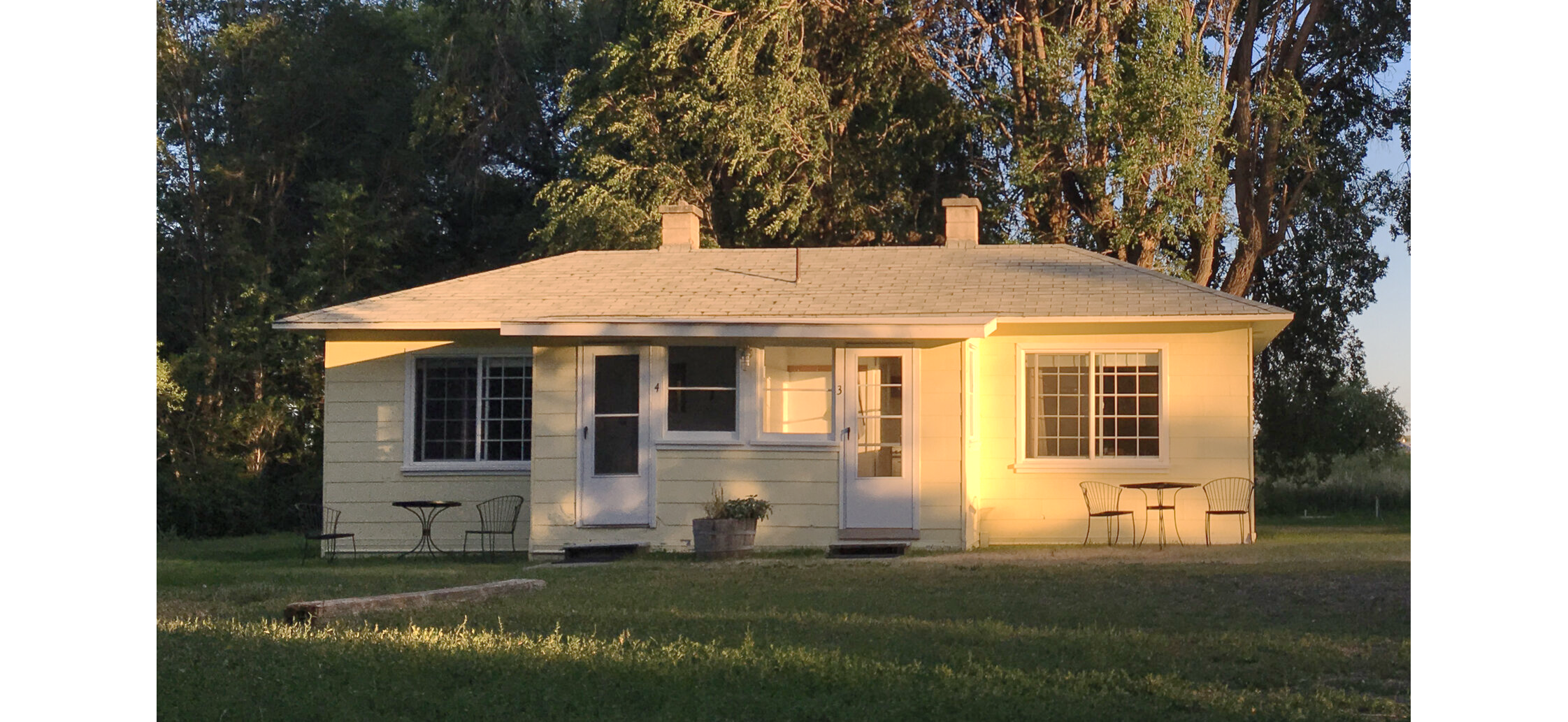 Photo of white single-story duplex cabin with lawn in front and trees behind, and golden sunset light reflecting vertically off a pipe.