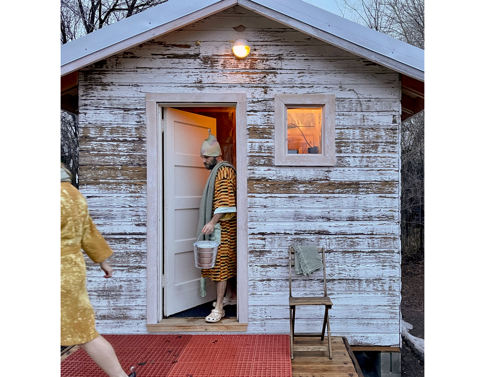 Photo of a man in a multi-colored, horizontally-striped bathrobe and white cap exiting the sauna and carrying a bucket of water.