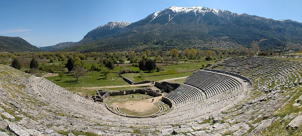 Photo of ancient Greek amphitheater with green fields and snow-capped mountain in background